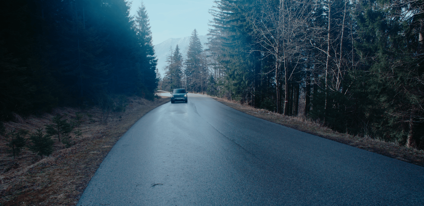 Ein Kia Fahrzeug fährt bei Dämmerung eine Waldstraße entlang und entfernt sich von der Kamera. Hohe Kiefern und Berge in der Ferne rahmen die Szene ein.