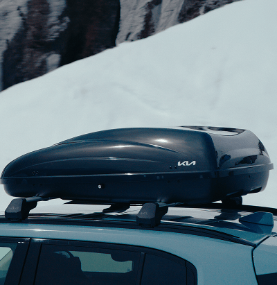 Kia Modell mit einer auf Dachträgern montierten Dachbox vor einer verschneiten Bergkulisse.