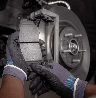 Nahaufnahme der behandschuhten Hände eines Technikers, der bei einer Inspektion einen Bremsbelag neben einer freiliegenden Bremsscheibe und einem Bremssattel hält.