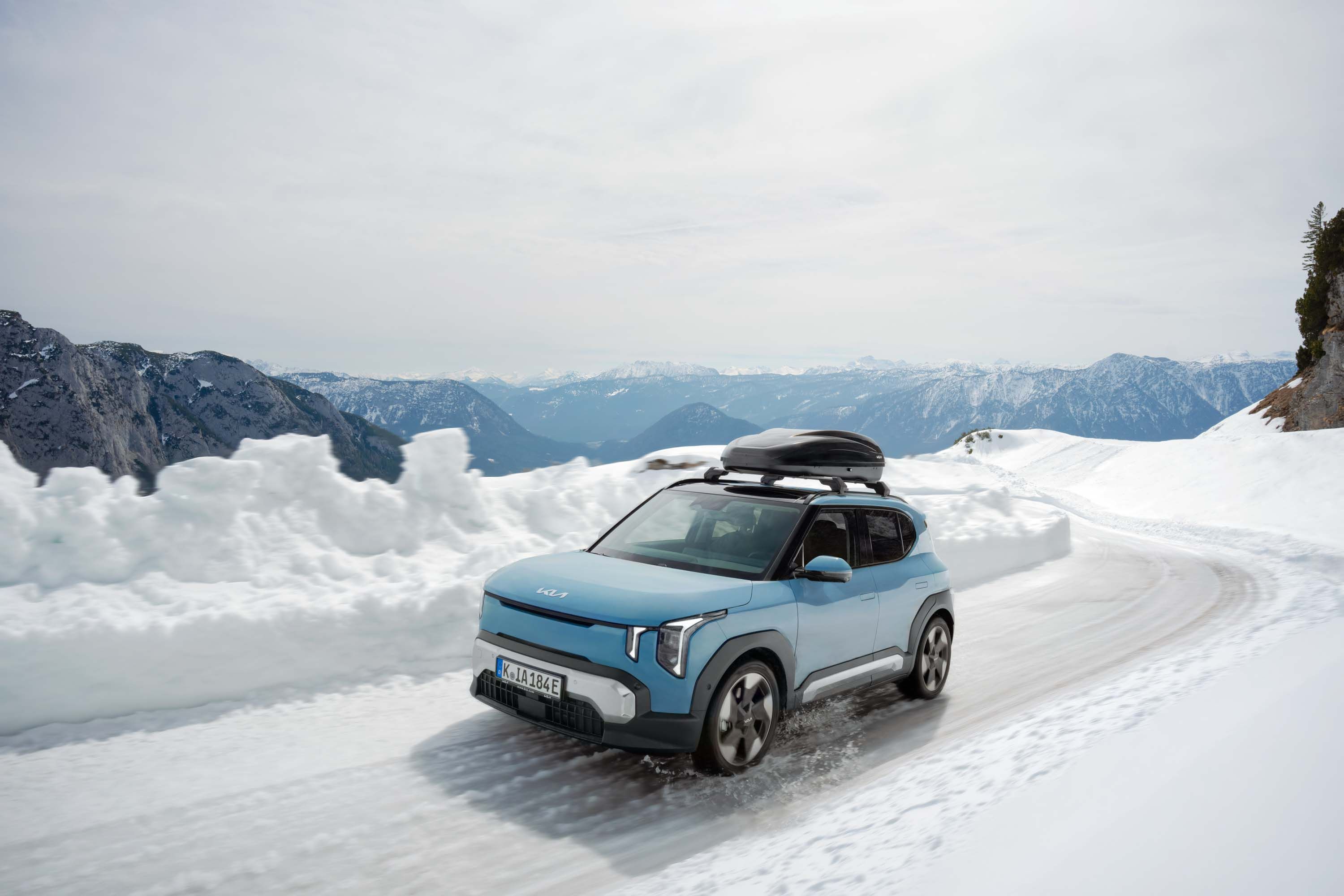Blue Kia SUV with a rooftop cargo box parked on a snowy mountain road, with a panoramic view of snow-capped alpine peaks in the background.