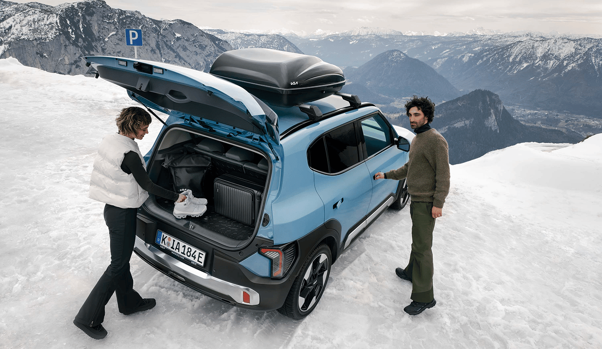 Two people loading luggage and shoes into the open trunk of a blue Kia SUV with a rooftop cargo box, parked on a snowy mountaintop with alpine scenery behind them.